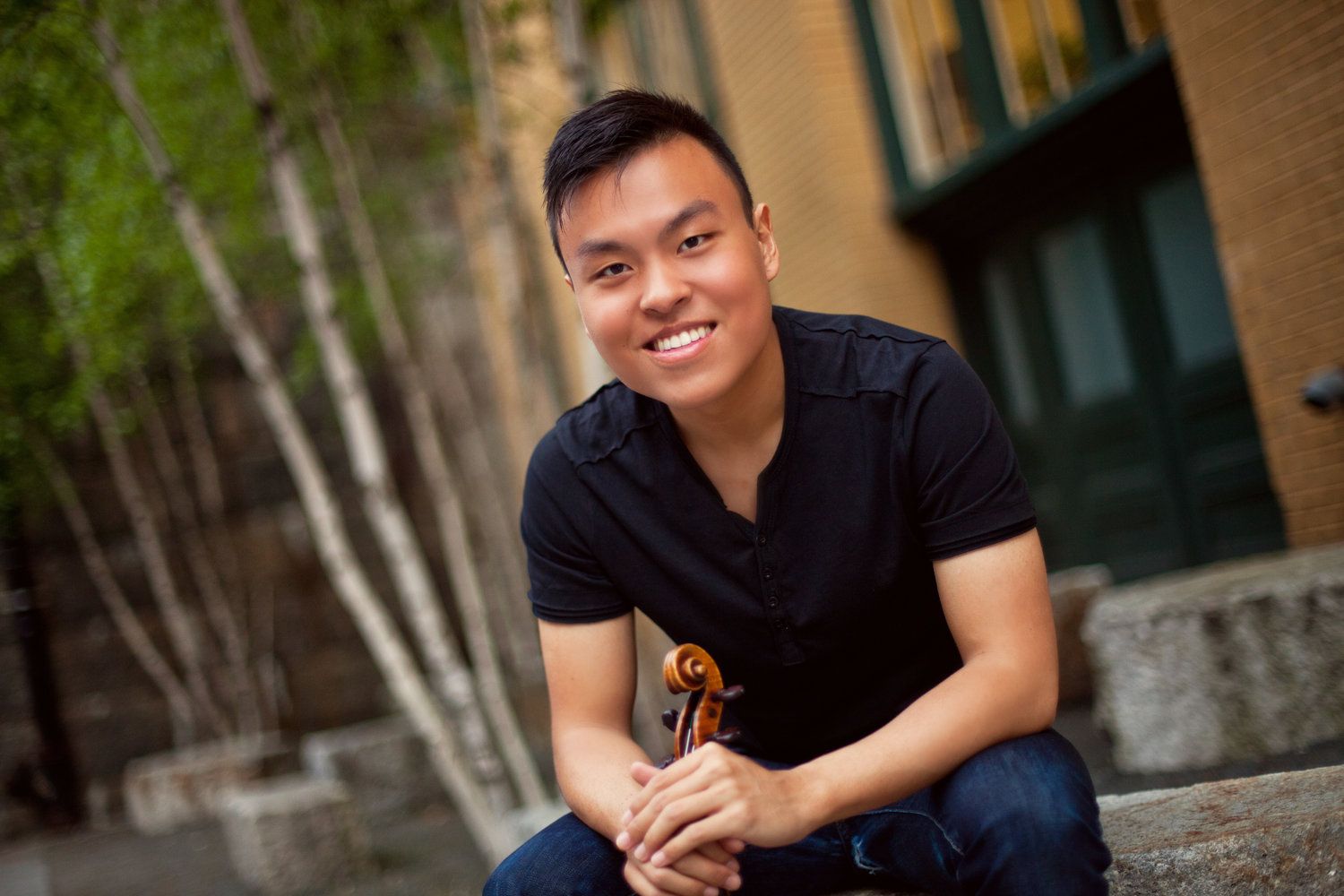 Luke Hsu smiles and holds his violin while sitting outdoors on some steps.