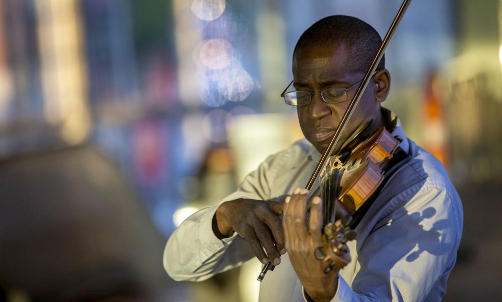 David France closes his eyes and plays his violin, with dappled evening light in the background.