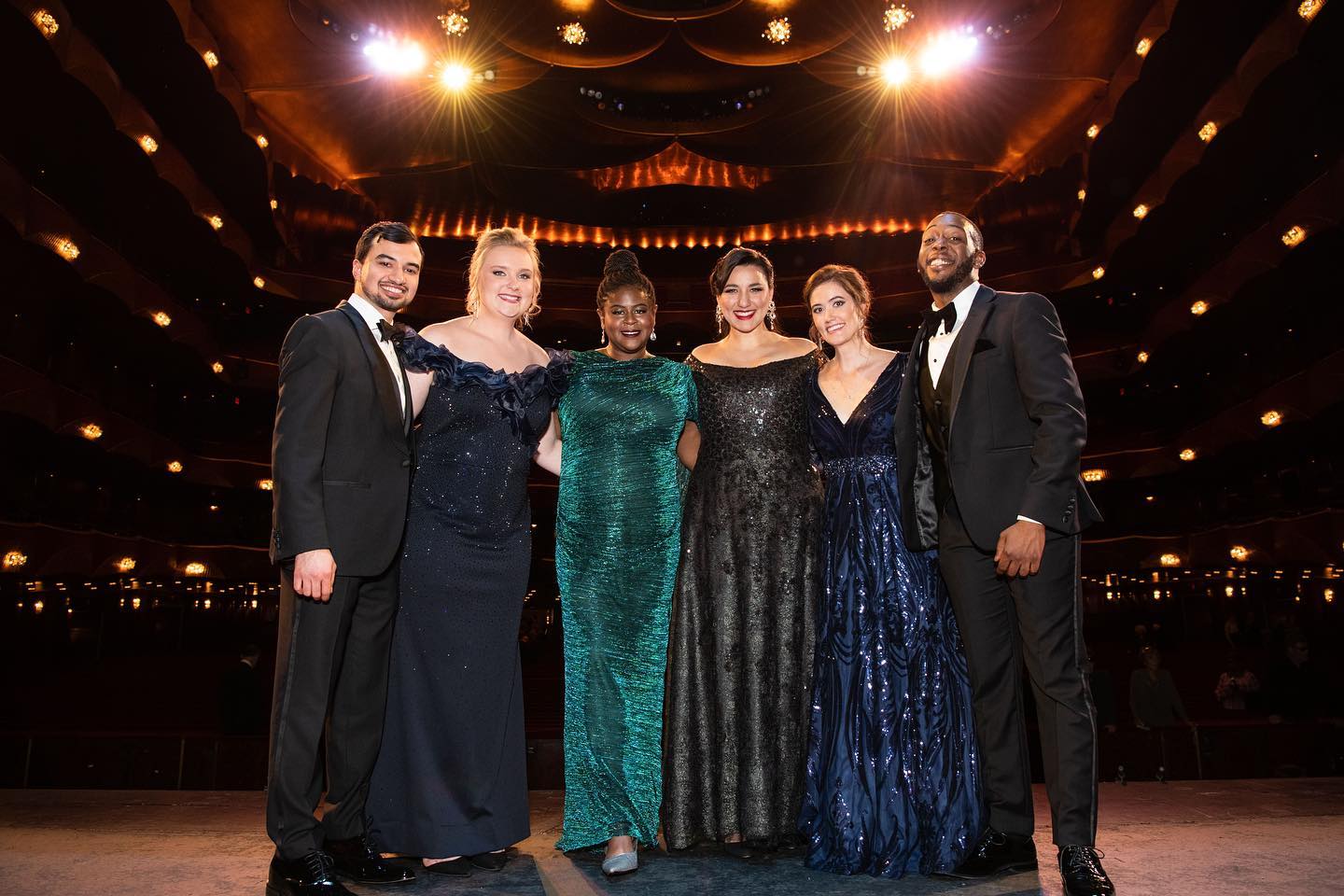 Six singers in tuxedos and gowns smile on the Met Opera stage