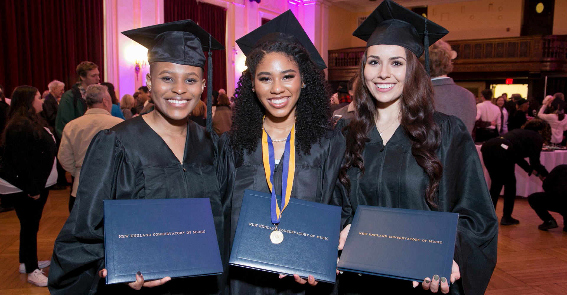 Three graduates smile and hold up their diplomas, wearing caps and gowns