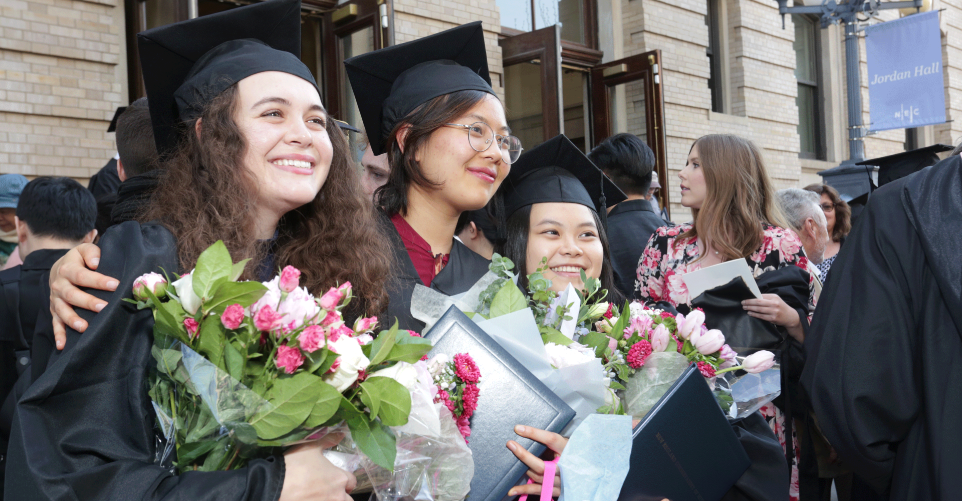 Students smile while wearing caps and gowns while holding bouquets in front of Jordan Hall