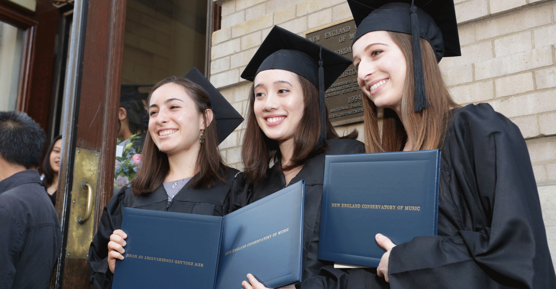 Three graduates smile and hold diplomas, standing on the steps of Jordan Hall