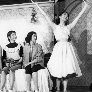 A scene from West Side Story, Maria smiles and opens her arms wide, while three women sit on a bed and look at her.
