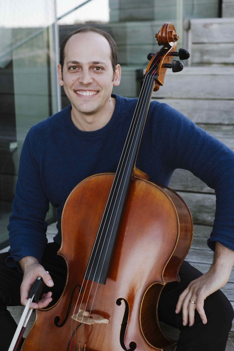 Mike Bloch smiles while wearing a blue shirt and sitting at his cello.