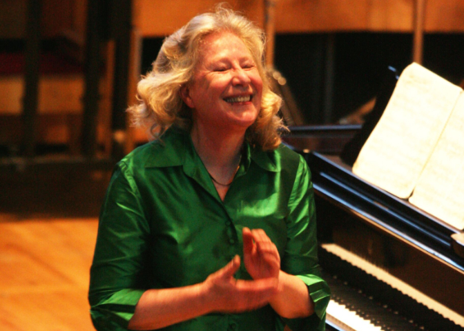 Ursula Oppens smiles and clasps her hands together with a piano in the background