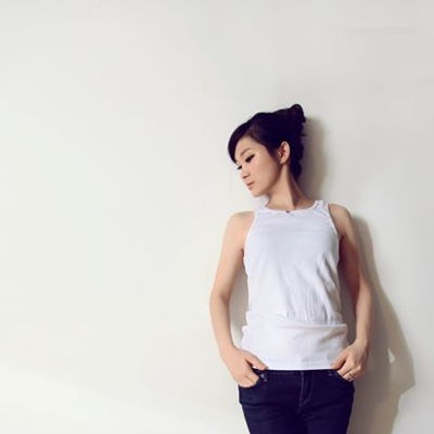 Angela Qianwen Shen leans against a white wall. She is looking away from the camera. Her hair is styled in an updo and her hands rest on her hips.