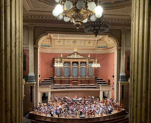 Wide shot of Youth Philharmonic orchestra playing in a grand concert hall with columns and a chandelier.