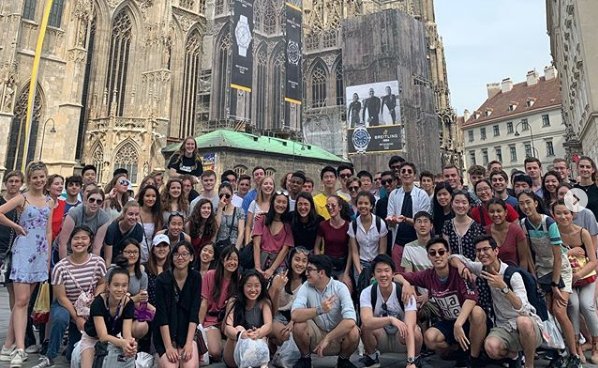 About 50 Youth Philharmonic members smile for the camera while standing in front of a historic building in Vienna.