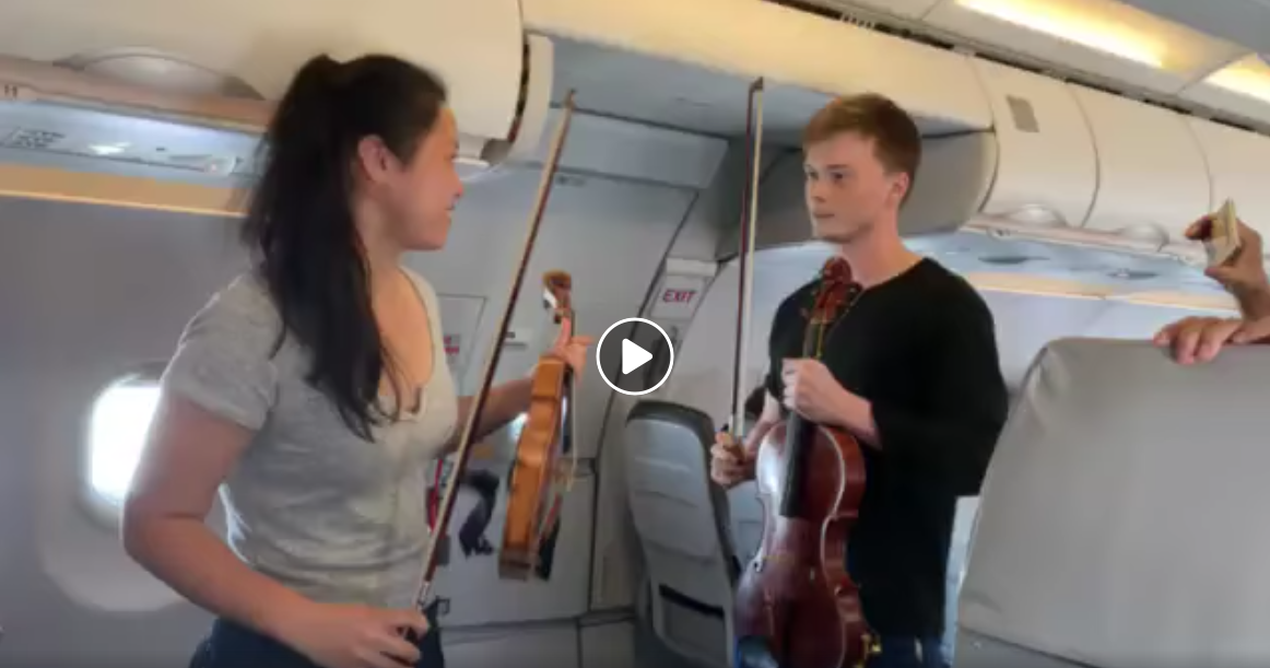 A violinist and violist stand in an airplane aisle with instruments in hand, about to play.