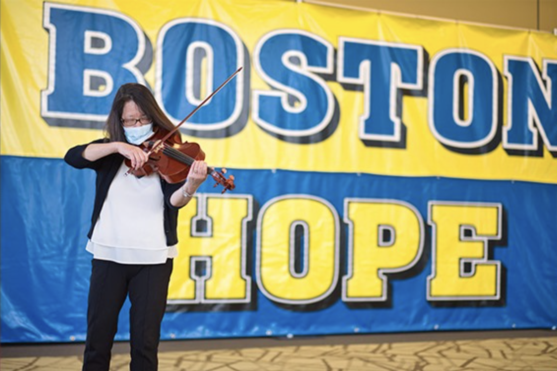 A violinist in a mask plays in front of a sign that reads Boston Hope