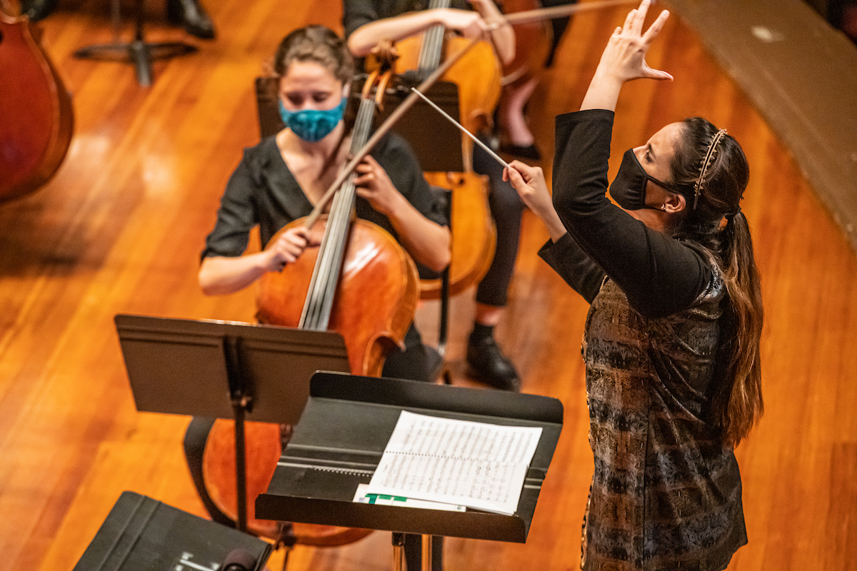 Lina Gonzalez-Granados tilts her head back while making an emotional gesture and conducting the NEC Philharmonia. Both Lina and the string players in the background are wearing masks.
