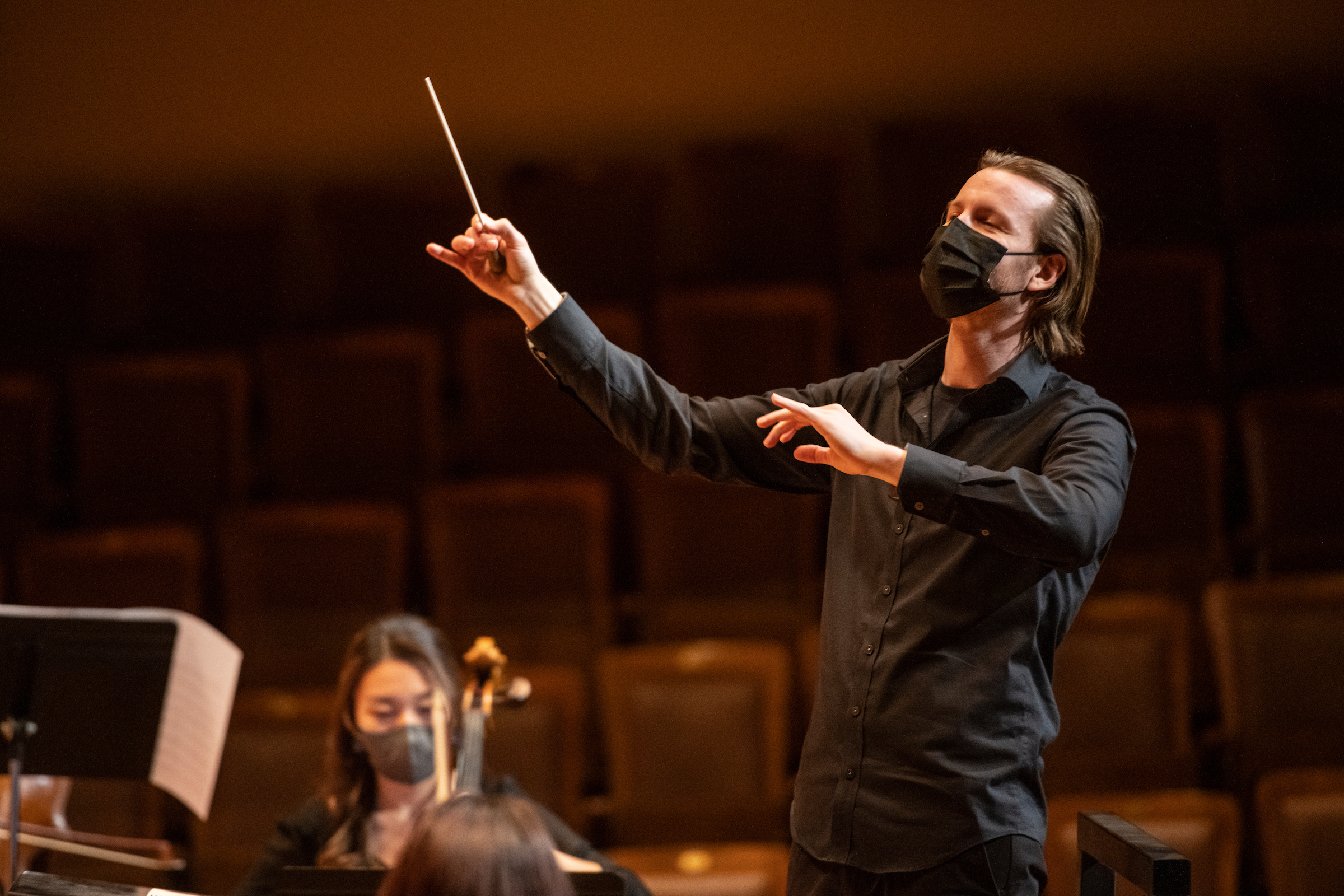 Tristan Rais-Sherman raises his baton while conducting the Philharmonia. He is wearing a mask and the empty seats of Jordan Hall are behind him.