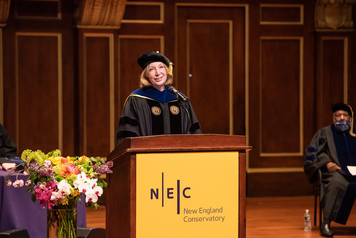 President Andrea Kalyn speaks from the Jordan Hall podium. She is smiling, and floral arrangements are on stage. She is wearing Commencement regalia.