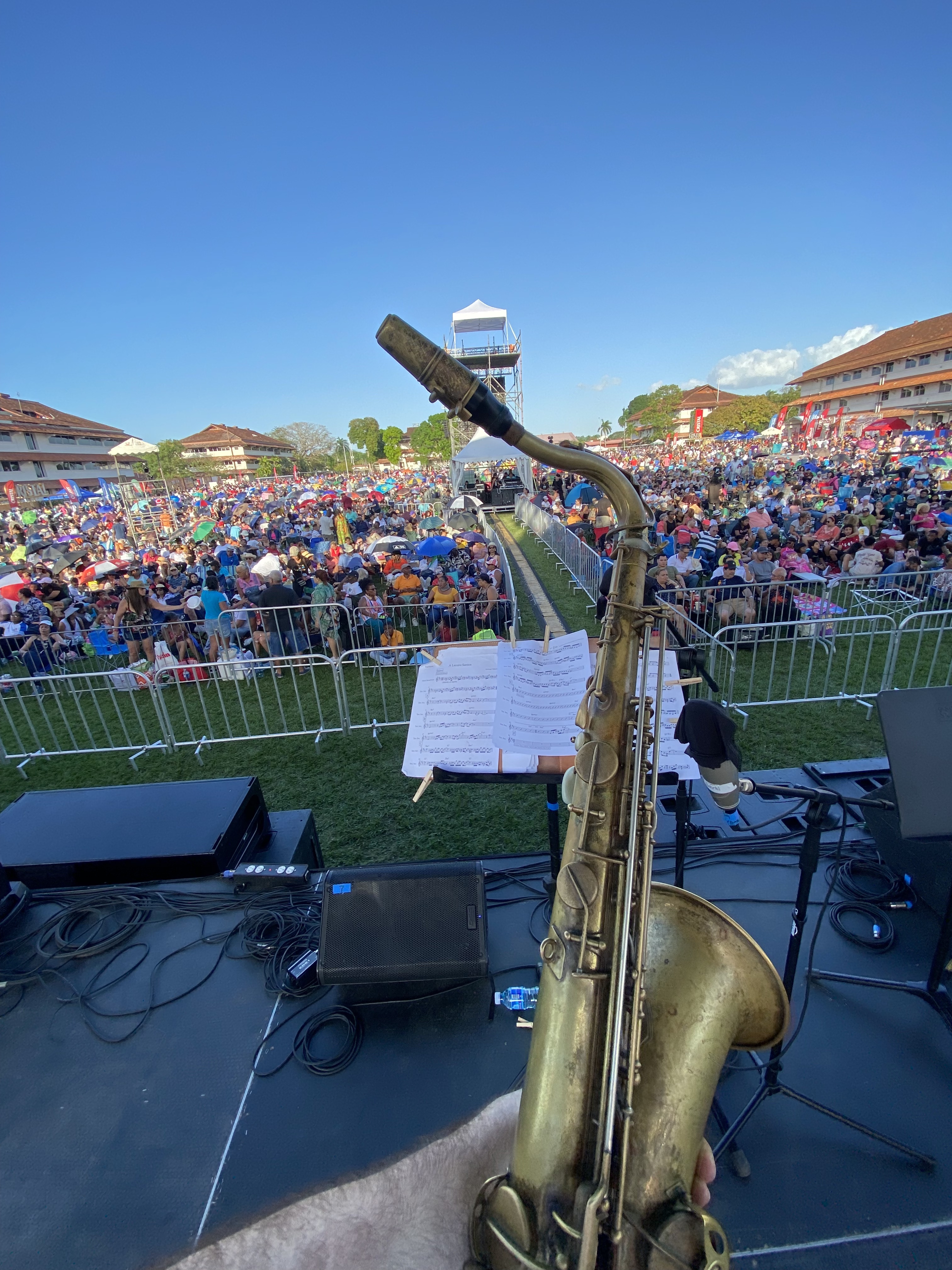 A jazz player holds his saxophone up in front of a large outdoor crowd at Panama Jazz Fest, with the blue sky in the background.