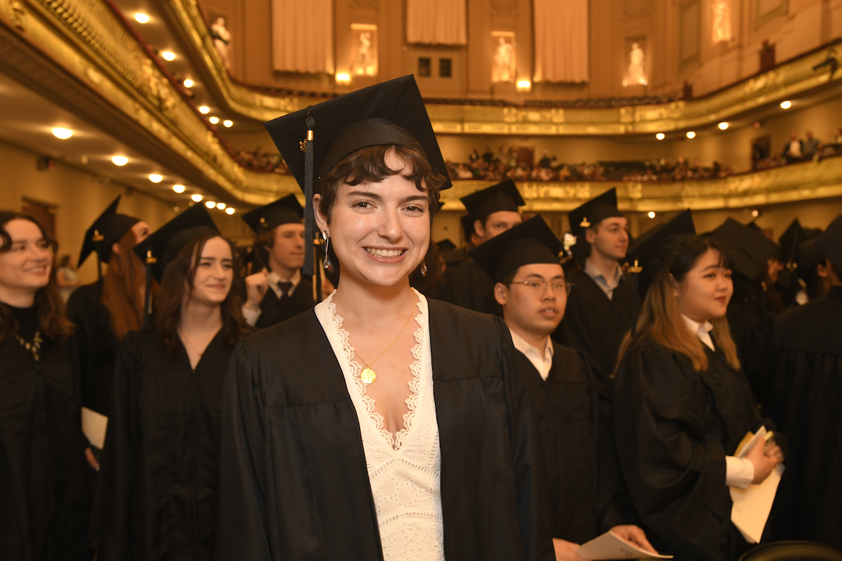 Brittany Bryant smiles with cap and gown in Symphony Hall, with other graduates behind her.