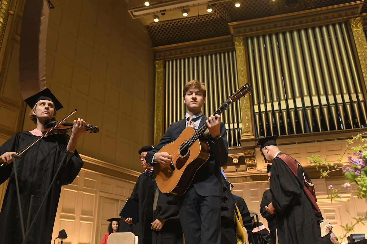 A fiddler in cap and gown and a guitarist in a suit play on Symphony Hall stage during graduation