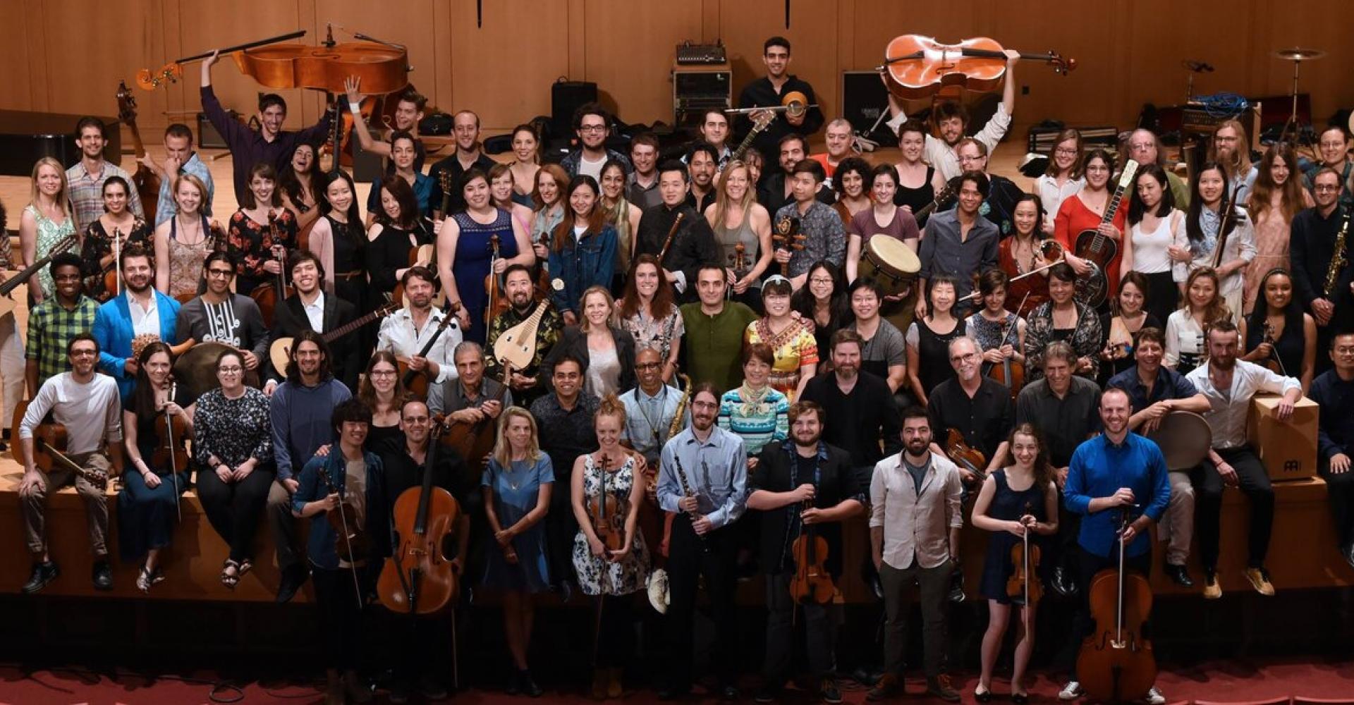 A large group of musicians from Silkroad's Global Musician Workshop crowd onto a stage to smile at the camera.
