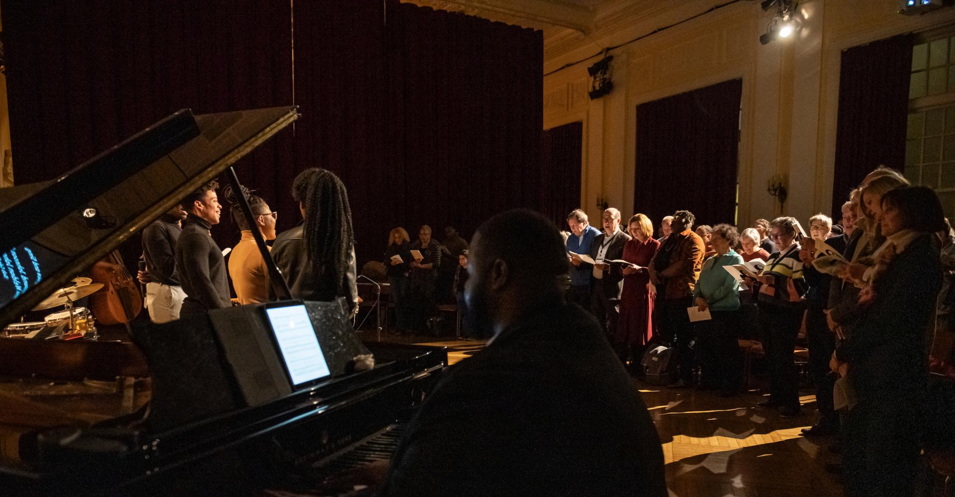 A pianist sits at the piano while students sing and the audience rises to sing with them.