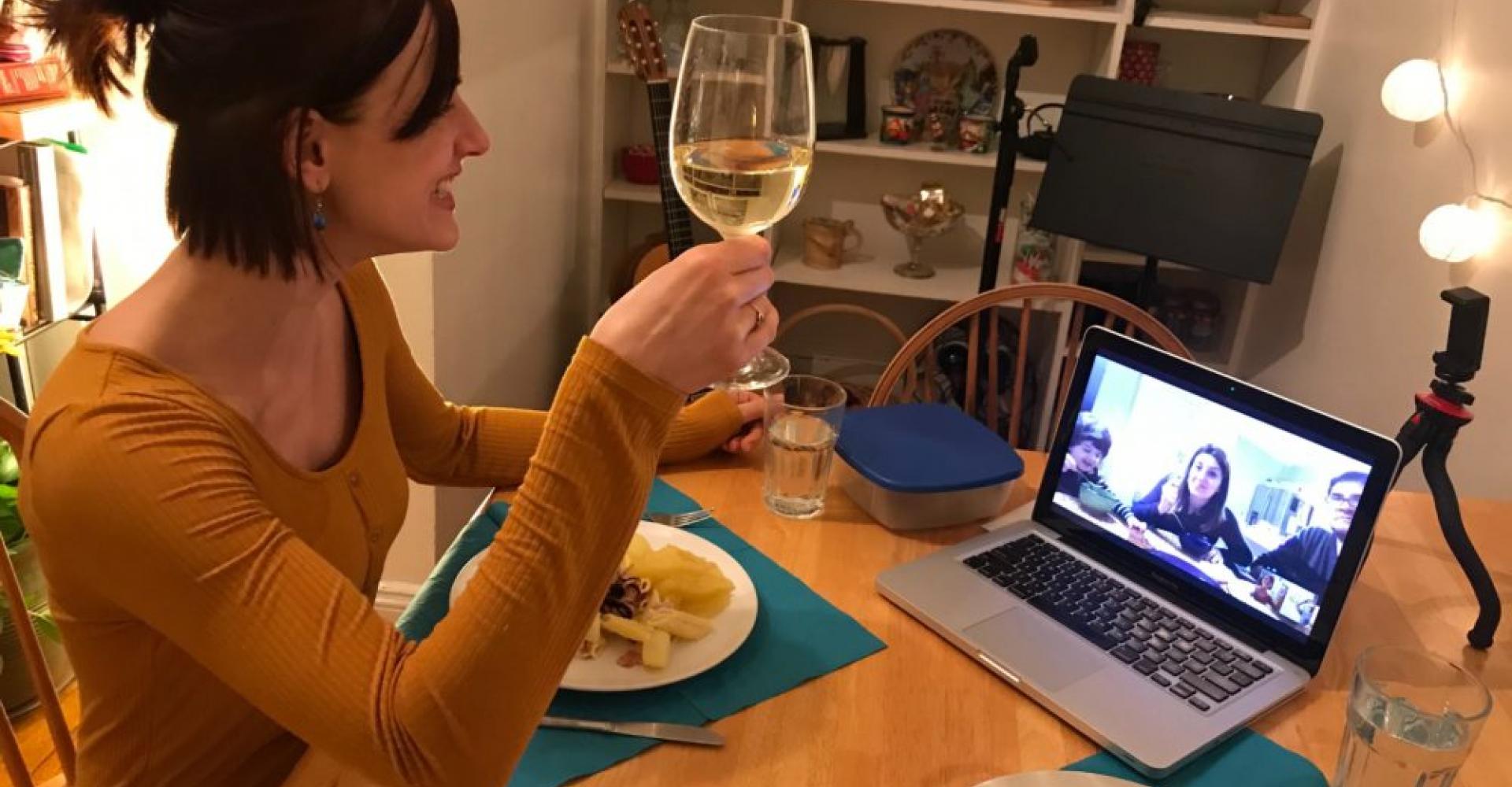 Gabriela Martina smiles and holds a wine glass while sitting down to dinner in her home. On screen is a fellow musician with whom she is virtually sharing the meal.