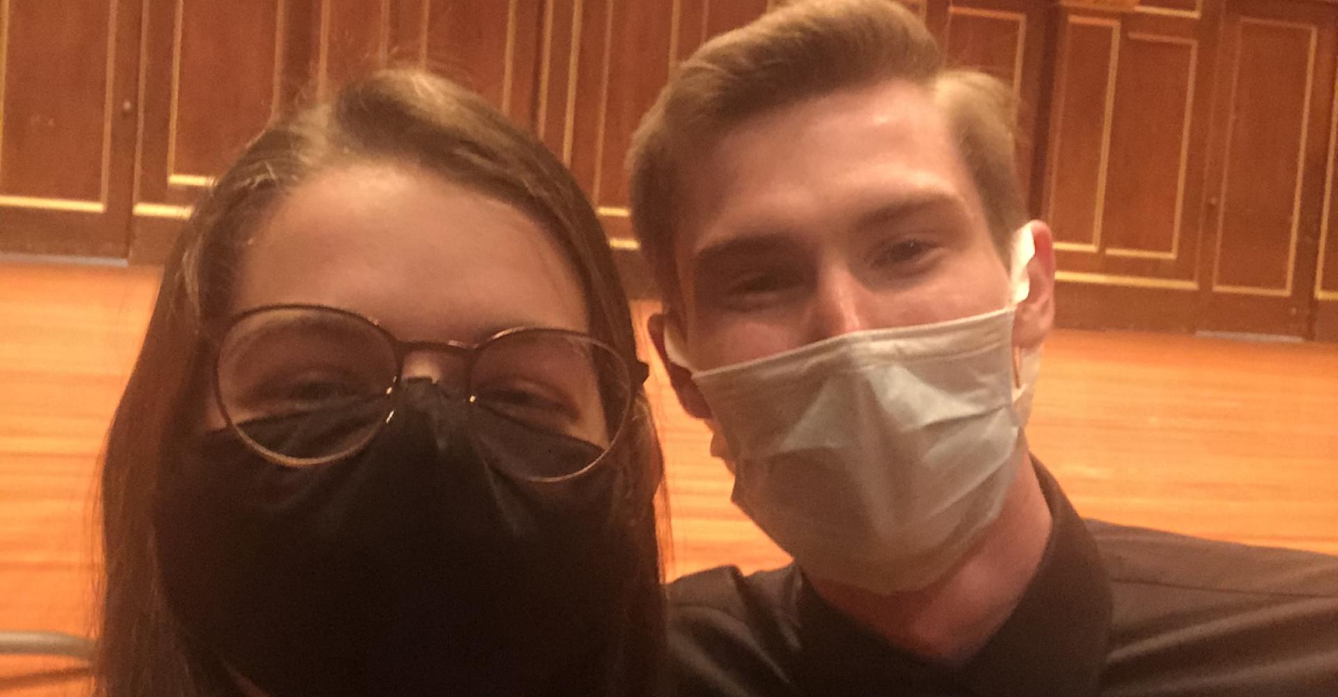 Jennie and Joseph take a selfie in Jordan Hall. Both are wearing concert blacks and masks, and the Jordan Hall stage is behind them.