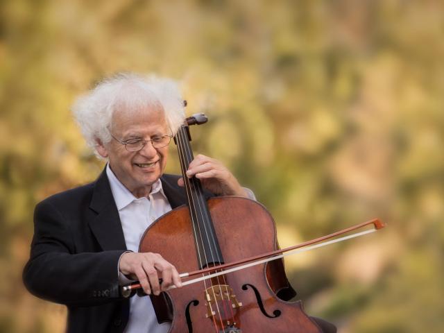 Laurence Lesser playing the cello and smiling, with green trees and plants in the background.