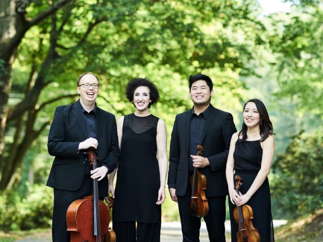 The Verona Quartet stands with their instruments under green-leafed trees.