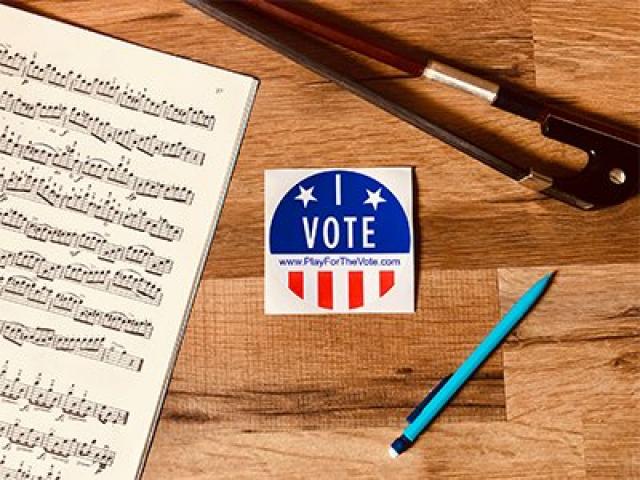An "I Vote" sticker on a desk with a cello bow, sheet music, and a pencil. The sticker also has the URL www.playforthevote.com