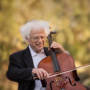 Laurence Lesser playing the cello and smiling, with green trees and plants in the background.
