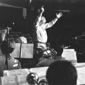 A smiling Gunther Schuller conducts an ensemble with his arms raised over his head, in a photo from 1990.