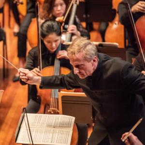 Conductor Hugh Wolff leans forward with an intense expression on his face, holding his conductor's baton. In the background, NEC Philharmonia string players perform.