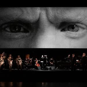 Performance shot of a video screen with eyes, projected above a chamber ensemble and chorus.