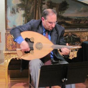 Mal Barsamian plays the oud while seated in front of a painting of trees.