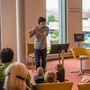 A flautist smiles and plays for an audience of young children, who raise their hands.
