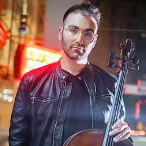 ID: Evan Kahn stands with his Cello in front of a blurred city street. He wears a black leather jacket