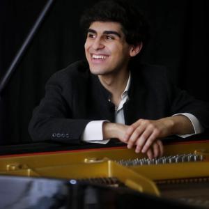 ID: Evren Ozel sits by an open piano, he faces to the left and is smiling.