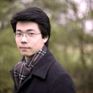 ID: Kuok-Wai Lio stands in front of a blurred natural background. He wears a scarf and jacket