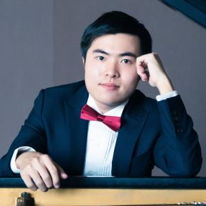 Zhiye Lin sits in front of a piano facing the camera. He wrests his head on his hand and is in formal attire.