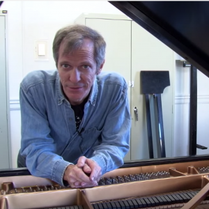 Steve Drury leans on a prepared grand piano looking at the camera