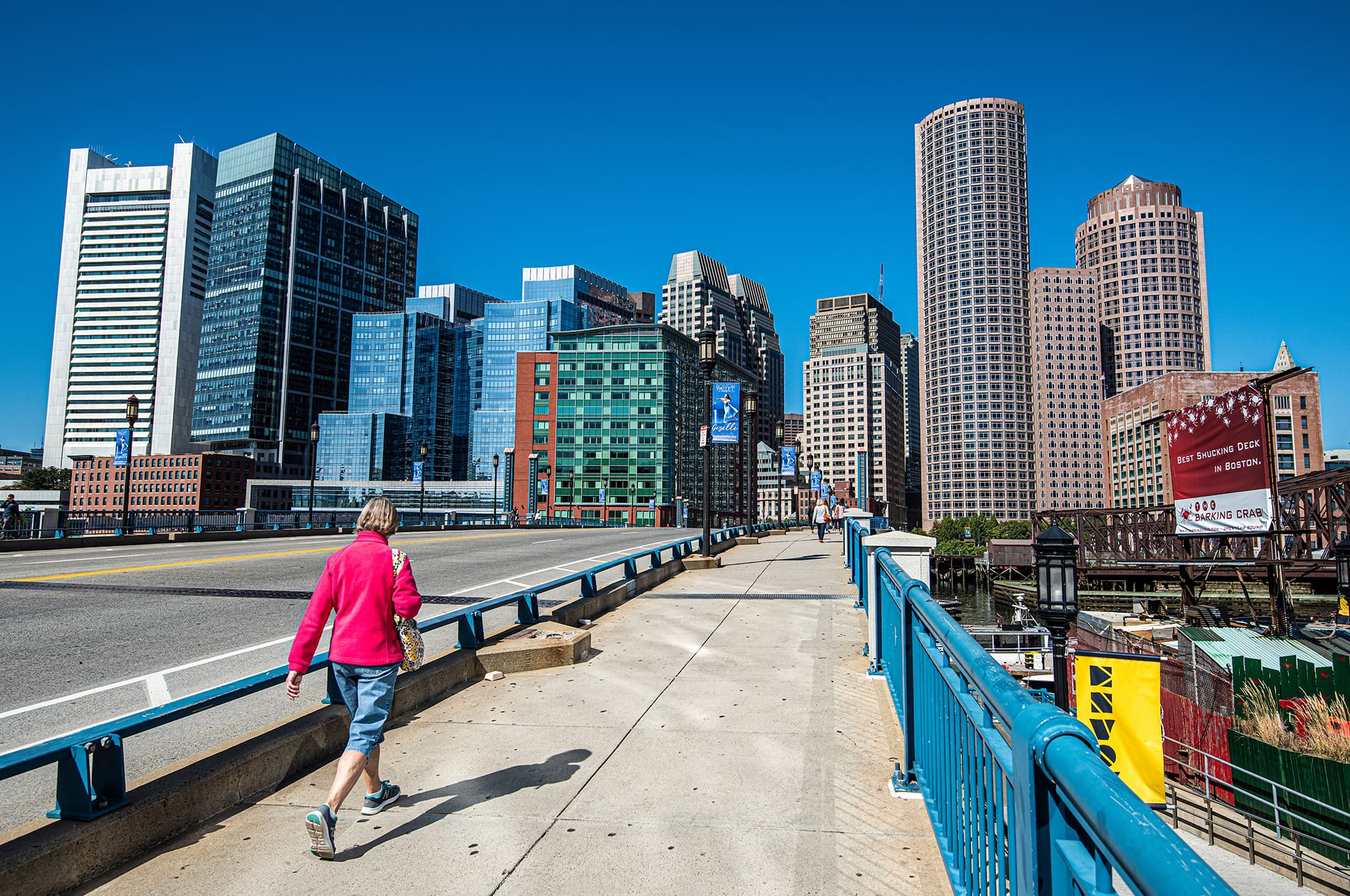 A person walking along a bridge in front of the Boston skyline.