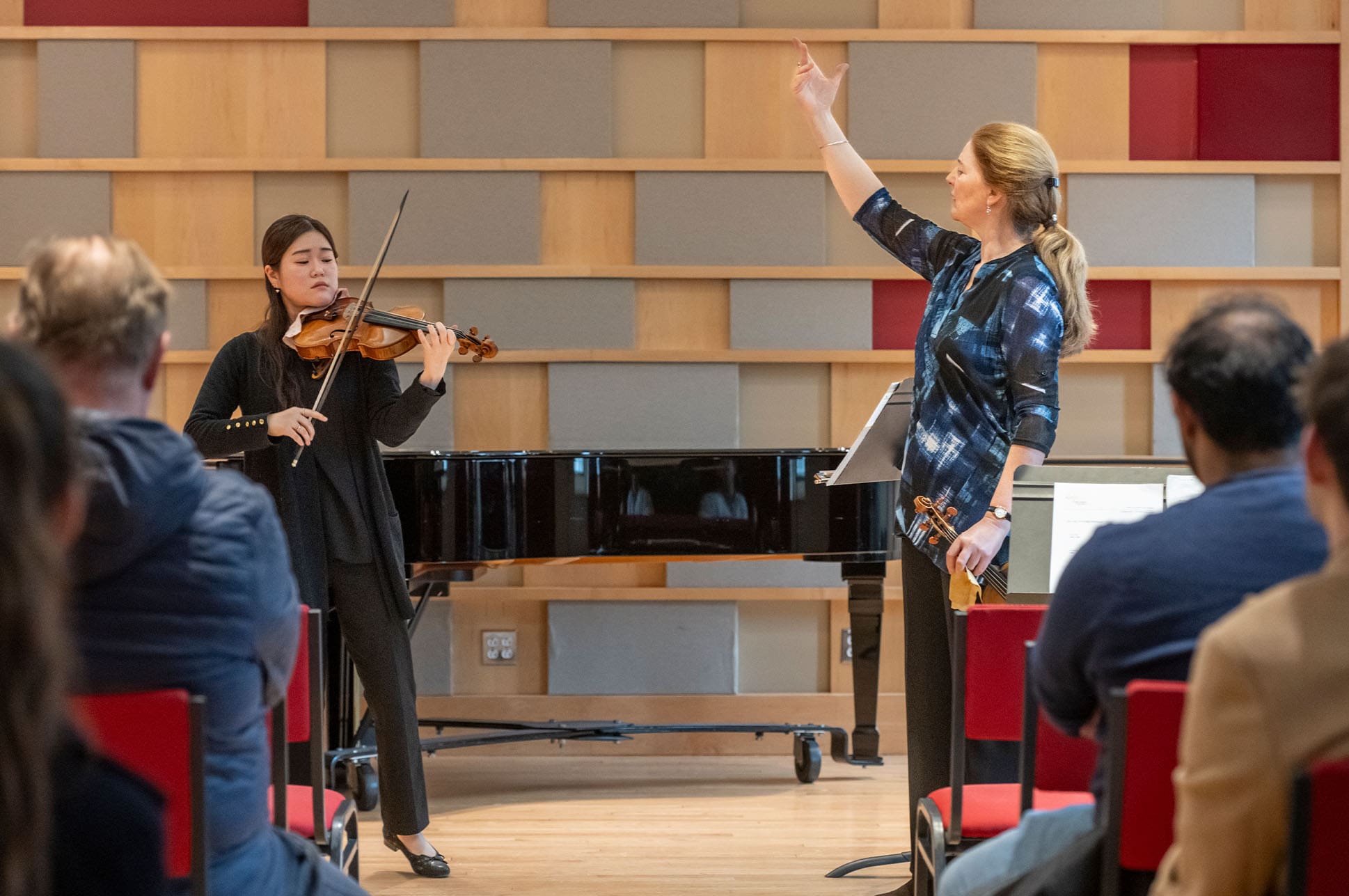 A student performing on violin in front of a classroom with an instructor guiding her.