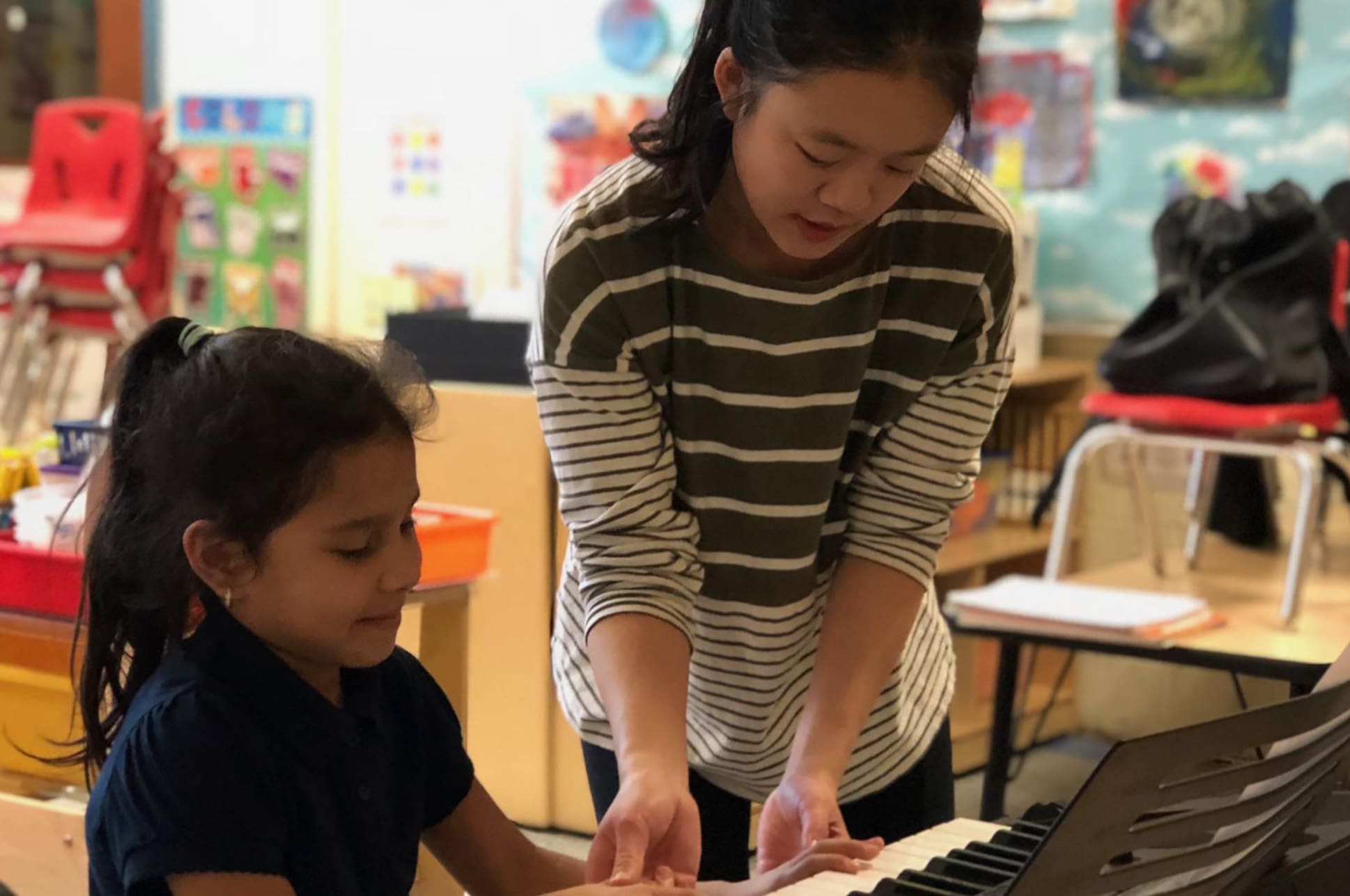 A person teaching a young child to play piano in a classroom.