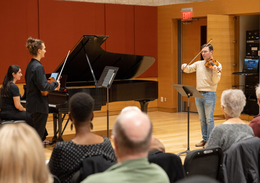 Student playing violin while another student plays piano, being instructed by a teacher.