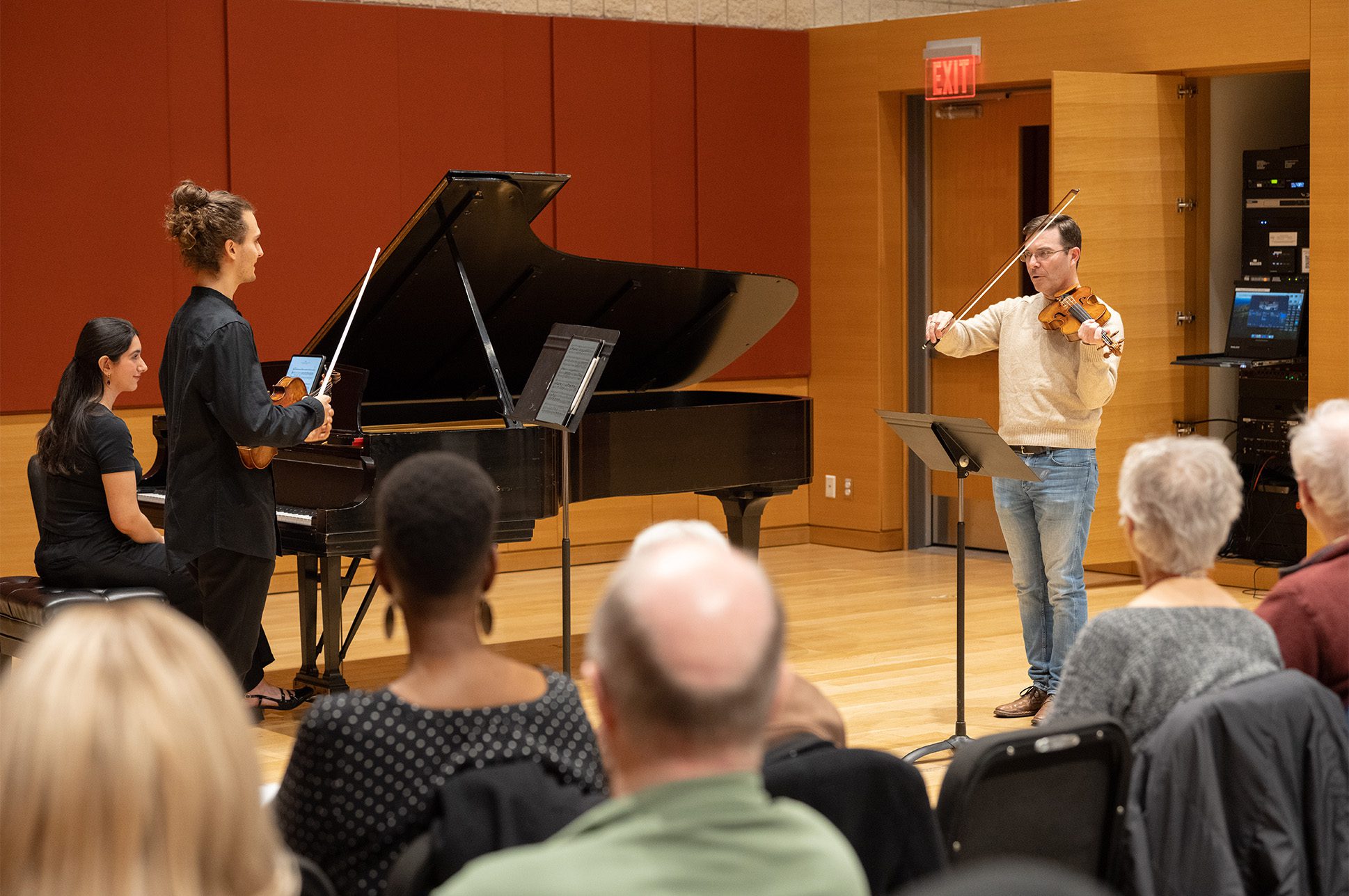 Student playing violin while another student plays piano, being instructed by a teacher.