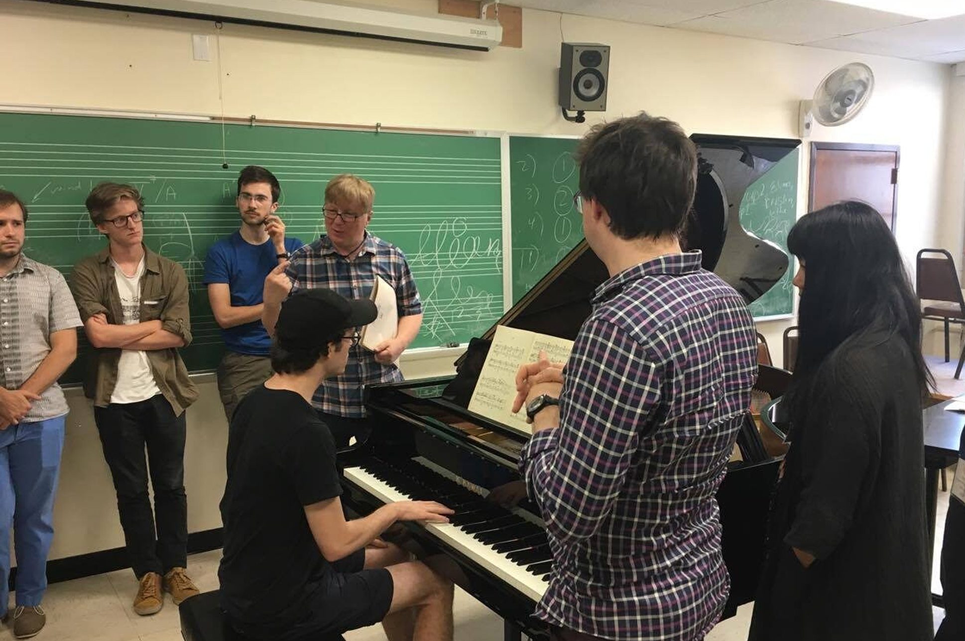 Students in a classroom surrounding an instructor on a piano. Students in a classroom surrounding an instructor on a piano.