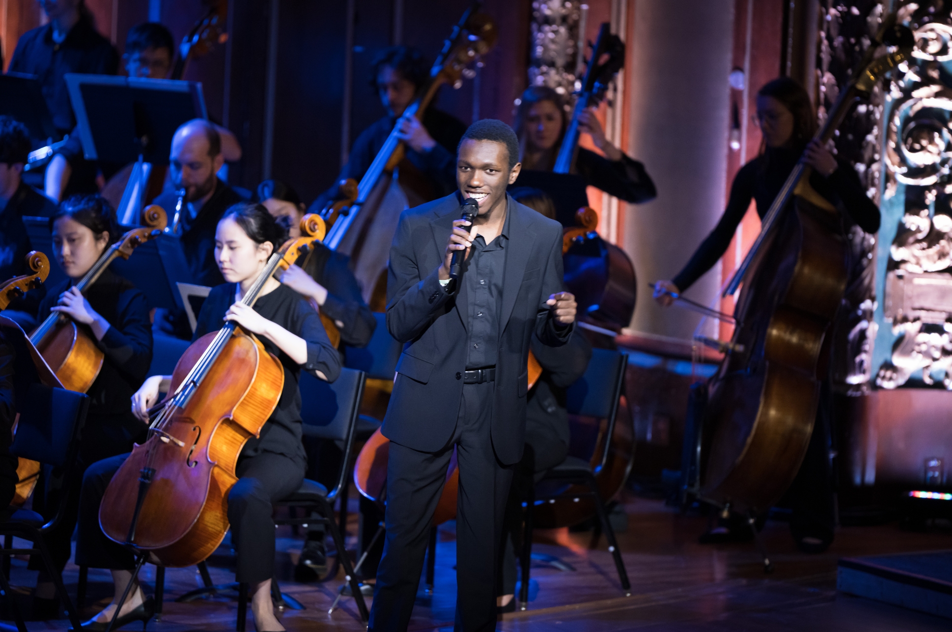 Student singing on stage with string players in background.